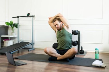 Atletik bir adam kişisel egzersiz sahasında bir dizüstü bilgisayarda çevrimiçi fitness kursuna katılırken minder üzerinde yan esneme egzersizi yapıyor.