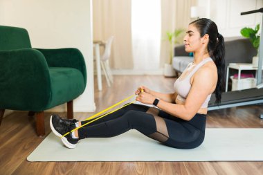 Genç bir kadın oturma odasında bacaklarını çalıştırırken direniş bandı kullanıyor.