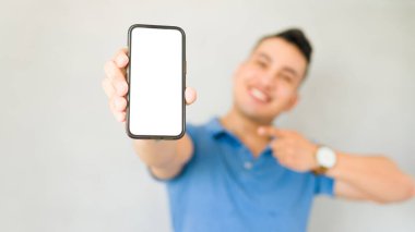 Gülümseyen adam, bulanık stüdyo arka planına karşı boş ekranla akıllı telefonu işaret ediyor.
