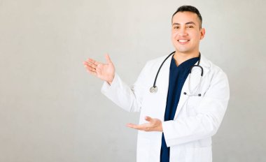 Erkek doktor kameranın önünde birine gülümserken elleriyle el hareketi yapıyor.