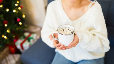 Beyaz süveter giyen bir kadın Noel ağacının önünde içinde lokum olan bir fincan sıcak çikolata tutuyor.