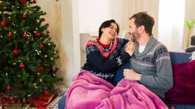 Mutlu çift kanepede battaniyenin altında oturmuş gülüyor ve Noel tatilinin tadını çıkarıyor.