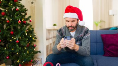 Adam arka planda süslenmiş bir Noel ağacı olan oturma odasındaki koltukta otururken akıllı telefon kullanıyor.