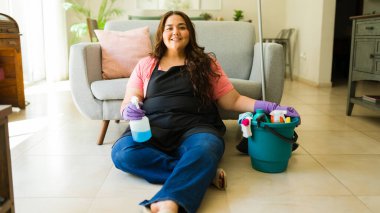 Ev işlerine ara verirken elinde sprey şişeyle yerde oturan mutlu temizlikçi kadın.