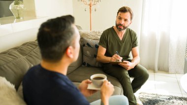 Yakışıklı Latin terapist kanepede kahve içen bir hastayla konuşurken not alıyor.