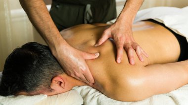 Bir masörün elleri, masaj masasında uzanmış bir adama sırt masajı yapıyor.