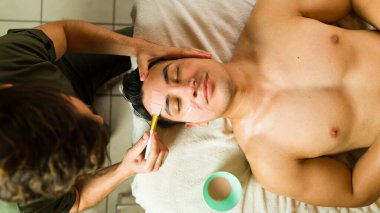 Erkek masöz, masaj masasında yatan genç bir adamın yüzüne kilden bir maske takıyor.