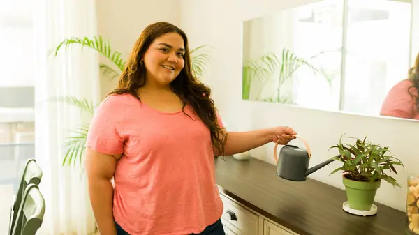 stock image Happy woman watering her houseplant at home, smiling for the camera. She's plus size, latin, in a cozy mexican apartment. Casual and content, symbolizing growth