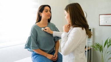 Doktor bir kadın hastanın kalbini dinlerken bir muayenehanede göğsüne steteskop dayıyor.