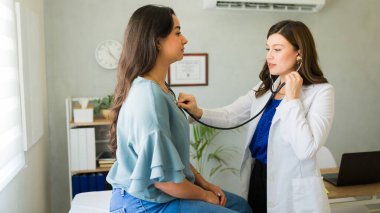 Kadın doktor, muayenehanesinde bir kadın hastanın nefesini dinliyor.