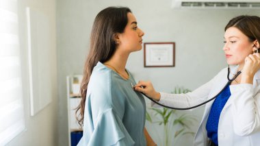 Kadın doktor, muayenesi sırasında steteskop kullanan bir kadının kalp atışlarını dinliyor.