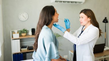 Genç bir kadın klinikte tıbbi danışmanlık yaparken boğazını bir kadın doktora muayene ettiriyor.