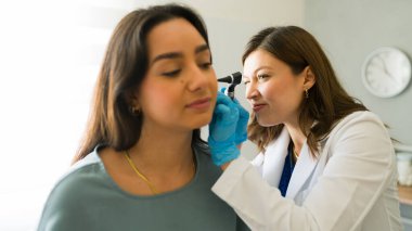 Laboratuvar önlüğü giyen kadın doktor genç bir kadın hastanın kulağını incelemek için otoskop kullanıyor.