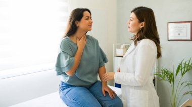 Latin bir kadın muayene masasında oturuyor ve bir doktora belirtilerini anlatırken boğazını tutuyor.