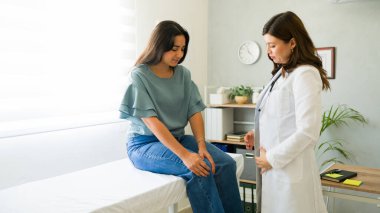 Doktor, genç bir bayanın muayene masasında otururken diz ağrısından bahsetmesini dinliyor.