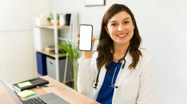 Tıp bürosundaki masasında boş bir ekranla akıllı telefonu tutarken gülümseyen kadın doktor.