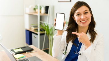 Kadın doktor, bir tıp bürosunda masasında otururken boş bir ekranla akıllı telefonu göstererek mutlu bir şekilde işaret ediyor.