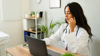 Kadın doktor ofisinde masasında oturuyor, laptopuyla çalışıyor ve telefonla konuşuyor.