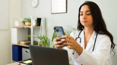 İspanyol kadın doktor ofisinde masasında oturuyor, akıllı telefon kullanıyor ve laptopuyla uğraşıyor.