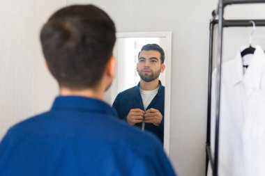 İyi görünümlü bir adam yatak odasındaki dolabın aynasında yansımasına bakarken gömleğinin düğmelerini ilikliyor.