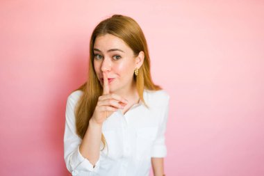 Uzun kızıl saçlı, beyaz gömlekli, işaret parmağını dudaklarında tutan tatlı bir kadın pembe arka planda sessizlik jesti yapıyor.