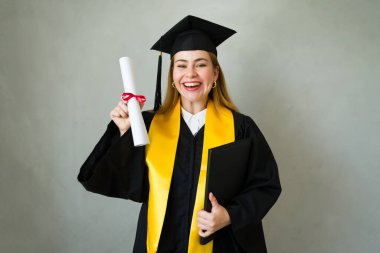 Şapka ve cübbe giyen genç mezun, diplomasını tutuyor ve stüdyo ortamında gülümsüyor.