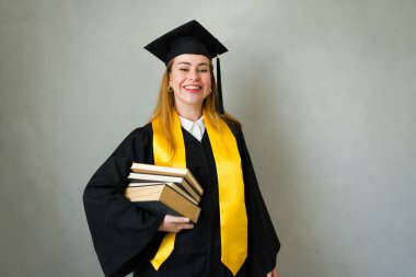 Güzel bir kadın üniversiteden mezun oluşunu kutluyor, kep ve cübbe giyiyor ve elinde bir yığın kitap tutuyor.