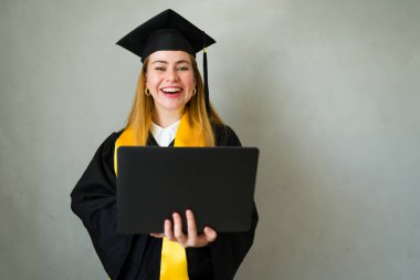 Genç mezun mezuniyetini resmen kutluyor, başlık ve cübbe giyiyor ve tören sırasında dizüstü bilgisayar tutuyor.