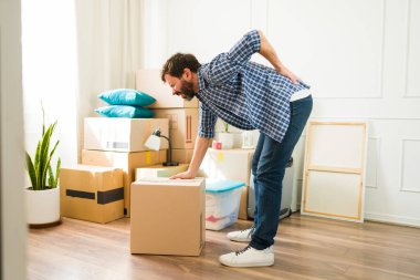 Yetişkin bir adam yeni dairesinde ağır bir kutuyu kaldırmaya çalışırken acı içinde kıvranıyor. Sırtını sıkıca tutuyor.