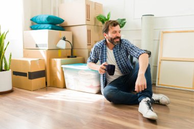 Gülümseyen adam kahve içiyor ve yeni evinin zemininde rahatlıyor. Etrafı kutularla çevrili.