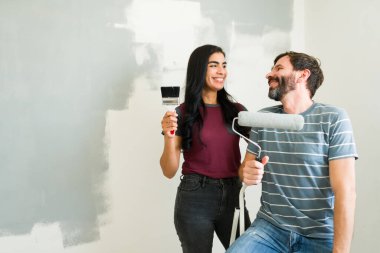 Genç çift gülümseyip birbirlerine bakarken ellerinde boya aletleri, yeni evlerinin duvarlarını boyamaya ara veriyorlar.