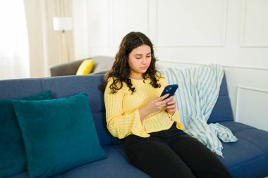 Latin genç kız evdeki oturma odasında rahat bir koltukta otururken cep telefonu kullanıyor.