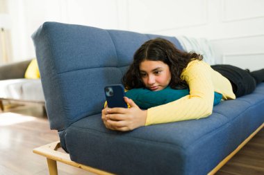 Latin kökenli gençler rahat ev ortamında koltukta dinleniyor, cep telefonu kullanıyor, sosyal medyanın ve online eğlencenin tadını çıkarıyor.