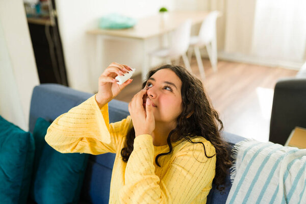 Teenager applying eye drops while sitting on a comfortable sofa in her living room, relieving dry or irritated eyes at home