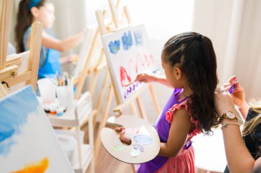 Tuval üzerinde resim yapan ve resim dersi sırasında elinde renkli resimlerle palet tutan sevimli bir anaokulu kızı.