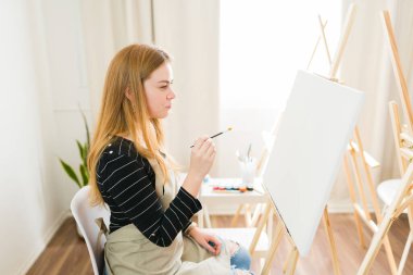 Genç bayan ressam, elinde fırçayla sanat eserlerine konsantre oluyor ve parlak bir sanat stüdyosundaki boş bir tuvale resim yapmaya başlamak üzere.