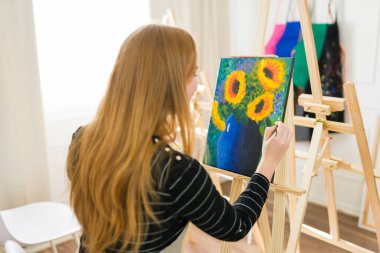 Dişi sanatçı, stüdyosunda boya fırçasıyla mavi bir vazoda ayçiçeği resmi yapıyor.