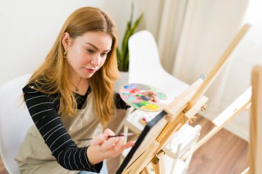 Genç bir kadın canlı bir sanat stüdyosunda palet ve fırçayla tutkuyla tuvale resim çizerek yaratıcı ruhunu ifade ediyor.