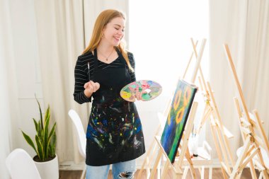 Resim fırçası ve paleti olan mutlu bir sanat eğitmeni resim dersinde bir sehpada ayçiçeklerini sergileyen bir tuval hayranı.