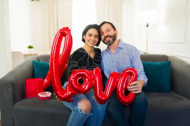 Kanepede oturan neşeli çift ellerinde kırmızı bir aşk balonu ve sevgililer gününü evde birlikte kutluyorlar.