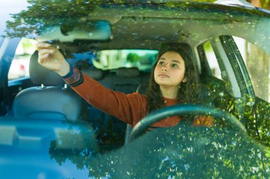 Güneşli bir günde araba sürerken arabanın dikiz aynasını ayarlayan genç bir kız.