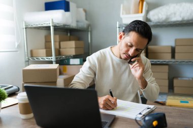 Latin girişimci bir müşteriyle telefonda konuşurken not alıyor ve küçük işletme dükkanında siparişleri yönetiyor.