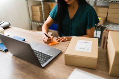 Laptop kullanırken ve depolama odasındaki ekomünikasyon siparişlerini yönetirken yapışkan bir nota notlar yazan etkili bir Latin girişimci.