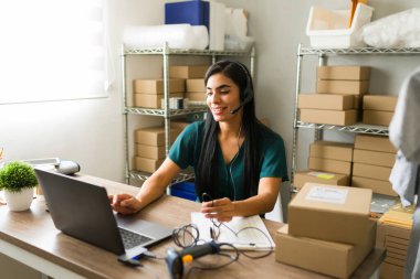 İspanyol girişimci kulaklık takıyor ve depo veya depolama odasında dizüstü bilgisayar kullanarak online sipariş ve teslimat yönetiyor.