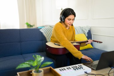 Dizüstü bilgisayarda çalışan, kulaklık takan, evde müzik bestelerken akustik gitar çalan bir bayan müzisyen.