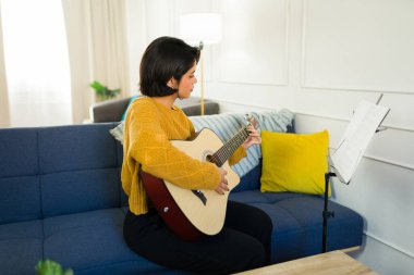 Oturma odasında rahat bir koltukta otururken akustik gitarıyla yeni bir şarkı öğrenen genç bir müzisyenin yan görüntüsü.