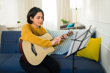 Konsantre kadın müzisyen akustik gitar çalıyor, nota kağıdını hazırlarken melodik yeterliliği geliştiriyor.