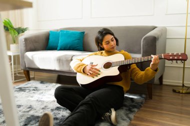Ahşap zeminde bacak bacak üstüne atmış, akustik gitar tıngırdatan ve evde melodik besteler yapan kadın müzisyen.