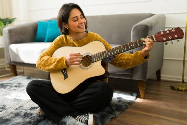 Gülümseyen İspanyol kadın akustik gitar performansından zevk alırken oturma odasında bağdaş kurup müzik tutkusunu ifade ediyor.