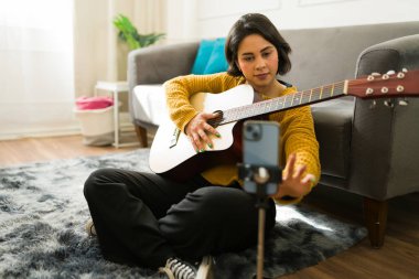 Güzel Latin kadın kendini kaydetmek için akıllı telefon kullanıyor. Oturma odasında gitar çalışıyor ve internetten müzik paylaşıyor.